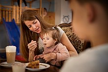 Eltern Kind Cafe Essen – Kaffee und Kuchen Eltern Kind Cafe | Kaffee & Kuchen – Mama gibt ihrer kleinen Tochter ein Stück Kuchen zu Essen mit einem Löffel. Auf dem Tisch steht ihr Latte Macchiato.