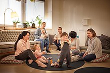 Kinderfreundliches Cafe Kinderfreundliches Cafe – Spielecke: Mamas sitzen auf Boden und spielen mit ihren Kindern und Babys. Ein kleines Kind lacht und zeigt in die Kamera.