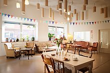 Das Anna Kindercafe: Leere Räumlichkeiten mit gemütlichen Sitzgelegenheiten, Sesseln, Tischen und Pflanzen. Sehr aufgeräumt, ordentlich und stilvoll eingerichtet. Von der Decke hängen dekorative Lampen und Lichte
