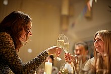 Event-Location Das Anna – Zwei junge Frauen stoßen lächelnd mit einem Sektglas an. Auf dem schön dekorierten Tisch brennt eine Kerze. Im Hintergrund sprechen auch andere Gäste mitienander.