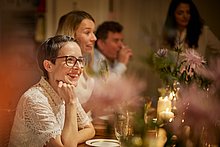 Event-Location: Frau sitzt am Tisch und unterhält sich lachend mit ihrem Gegenüber. Schön und feierlich dekorierter Tisch.