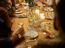 Event-Location Das Anna – Catering & exklusives Essen: Perspektive über die Schulter eines Gastes blickend auf den Tisch. Dort sieht man Hände, die Sektgläser halten, schön angerichtete Vorspeisen und feierliche Tischdekoration.