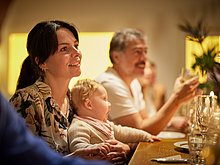 Event-Location: Frau sitzt mit Ihrem Baby auf dem Schoß am Tisch und schaut aufmerksam und lächelnd ihren Gesprächspartner auf der anderen Seite des Tisches an. Das Baby gähnt.