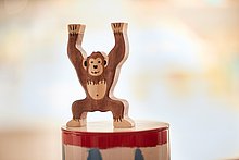 Das Anna Shop: Kleiner Spielzeugaffe aus Holz steht auf einer Dose und reckt die Arme lächelnd in die Höhe.